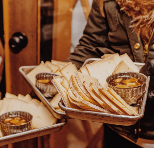 A person holds two trays of sliced cheese and bowls of dipping sauce, set against a rustic wooden background.
