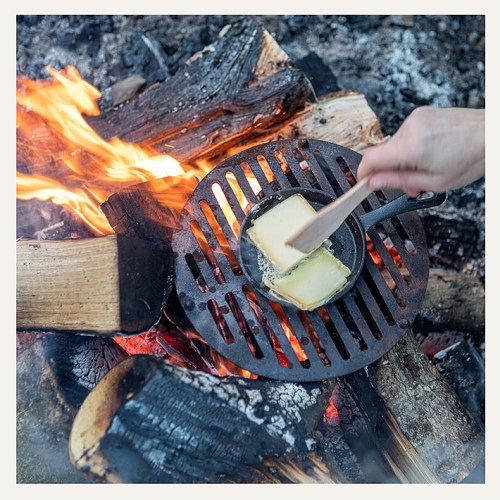 A hand stirs butter in a small skillet over a campfire, surrounded by burning logs and glowing embers.