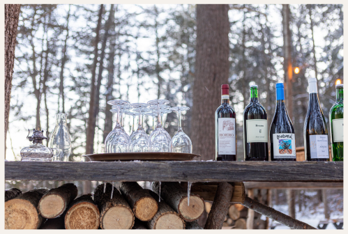 A rustic wooden table displays wine bottles and glasses, surrounded by snowy trees in a winter setting.