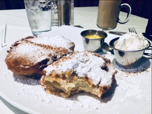 Two powdered sugar-dusted pastries on a plate, accompanied by a small cup of cream and a cup of yellow sauce.
