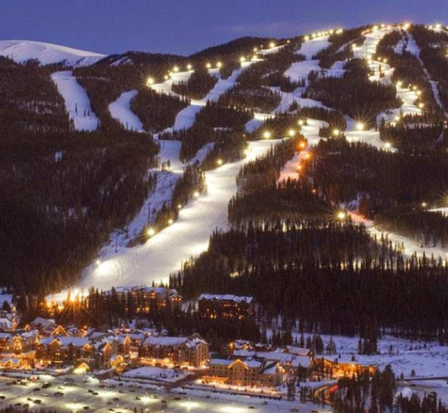 A snowy mountain landscape at dusk, with ski slopes illuminated by lights and a cozy village at the base.