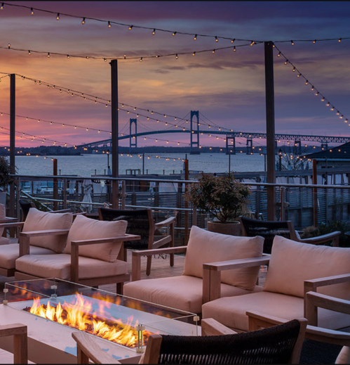 Cozy outdoor seating with a fire pit, overlooking a bridge at sunset, adorned with string lights.