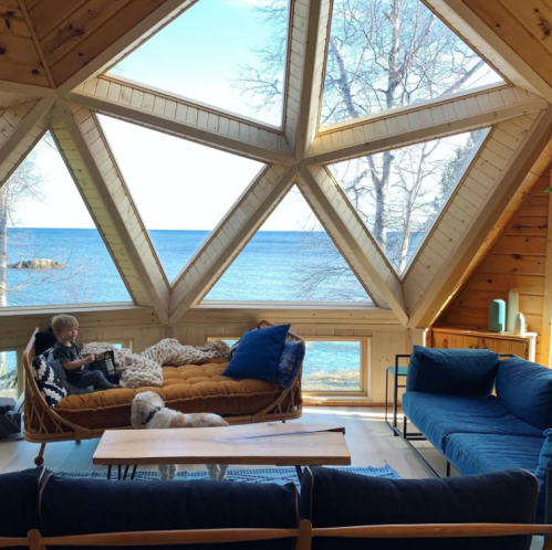A cozy, modern cabin interior with large windows overlooking a lake, featuring a person relaxing on a sofa and a dog nearby.