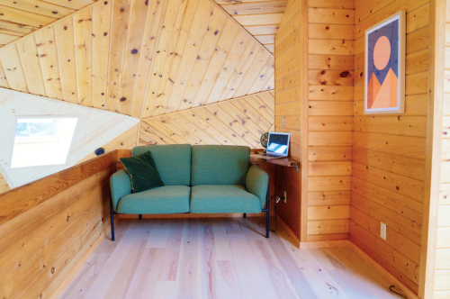 Cozy corner with a green sofa, wooden walls, and a small desk with a laptop, featuring a colorful wall art piece.