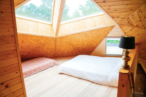 Cozy attic bedroom with wooden walls, skylights, and two beds, one with a patterned blanket and a lamp nearby.
