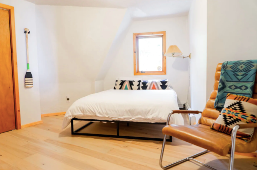 Cozy bedroom with a bed, wooden floor, a chair, and a window, featuring minimalist decor and warm lighting.