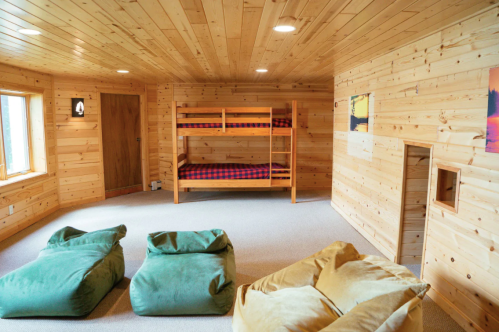 Cozy wooden room with a bunk bed, two bean bags, and large windows, featuring warm, natural lighting.