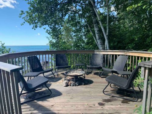 A wooden deck with six chairs surrounding a fire pit, overlooking a serene lake and lush greenery.