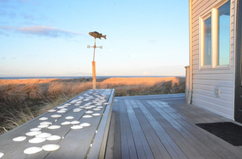 A wooden deck with white circular objects, a fish weather vane, and tall grass under a clear blue sky.