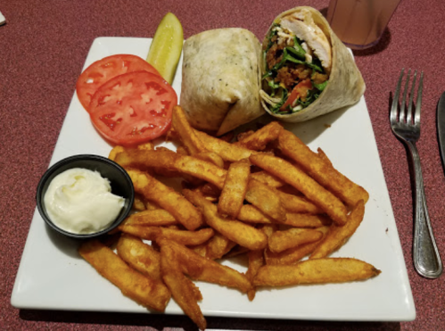 A plate with a wrap filled with vegetables, served with fries, tomato slices, a pickle, and a small cup of sauce.