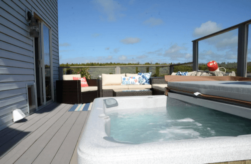 A hot tub on a deck with outdoor seating, surrounded by greenery and a clear blue sky.