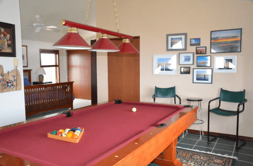 A pool table with a maroon felt surface, surrounded by chairs and framed photos on the wall in a bright room.