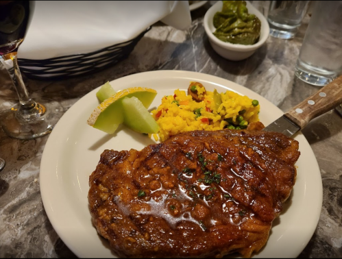 A plate featuring a glazed steak, yellow rice with vegetables, and melon slices, with a drink and condiments in the background.