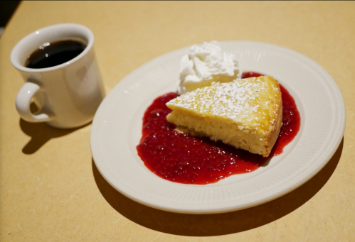 A slice of cheesecake topped with raspberry sauce and whipped cream, served with a cup of coffee on the side.