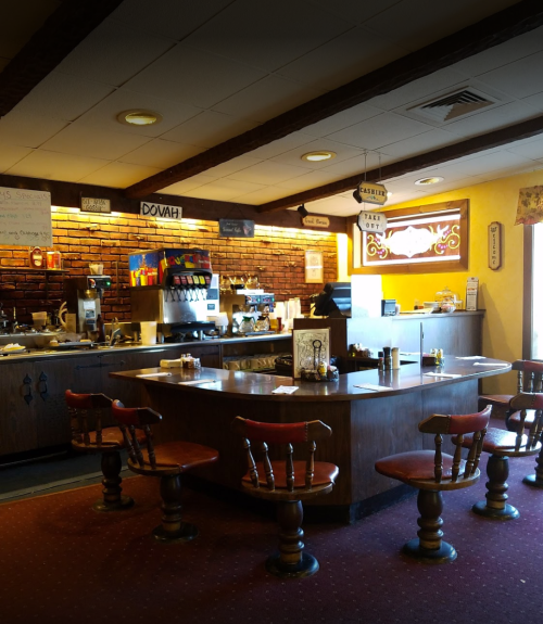 Cozy restaurant interior with wooden chairs, a brick wall, and a counter displaying drinks and condiments.