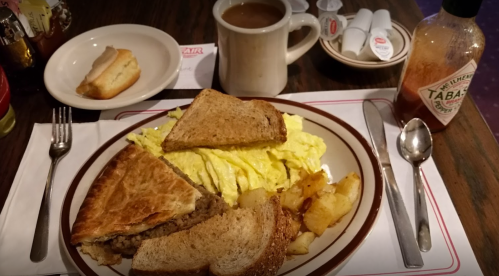 A plate with scrambled eggs, a sandwich, hash browns, a biscuit, and a cup of coffee on a table.