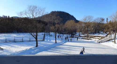 A winter scene with people ice skating on a frozen pond, surrounded by trees and a distant hill under a clear blue sky.