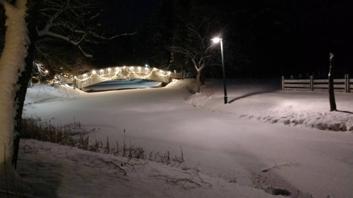 A snowy landscape at night featuring a bridge adorned with lights and a lamp post illuminating the scene.