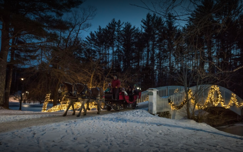 A horse-drawn sleigh passes over a snow-covered bridge adorned with festive lights at dusk.