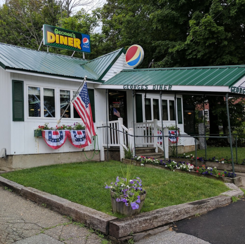 A charming diner with a green roof, American flags, and flower beds, featuring a Pepsi sign outside.