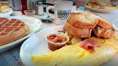 A plate of breakfast featuring an omelette, toast, baked beans, and a side of waffles, with a coffee cup in the background.