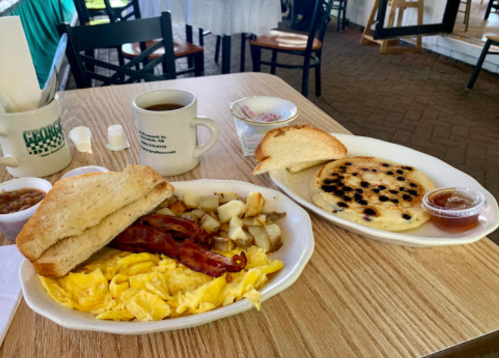 A breakfast spread featuring scrambled eggs, bacon, potatoes, toast, and a pancake with blueberries, alongside coffee.