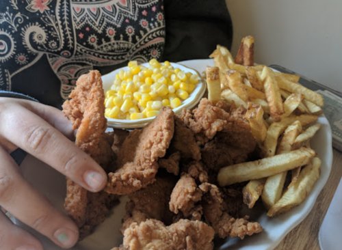 A plate of fried chicken, corn, and French fries, held by a person wearing a patterned shirt.