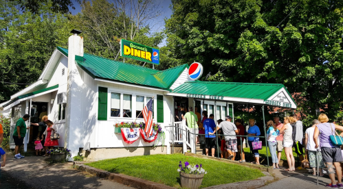 A busy diner with a green roof, people waiting in line outside, and patriotic decorations on the building.