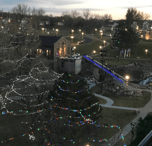 A park decorated with colorful lights, featuring trees, pathways, and a small building, all illuminated at dusk.