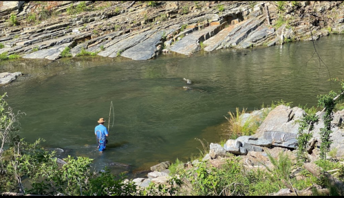 A person fishing in a calm river surrounded by rocky terrain and greenery.