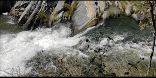 A rushing river flows over rocks, surrounded by greenery and a rocky landscape.