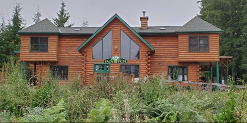 A large log cabin with a green trim, surrounded by tall grass and trees, featuring a sign that reads "Pace Pride."