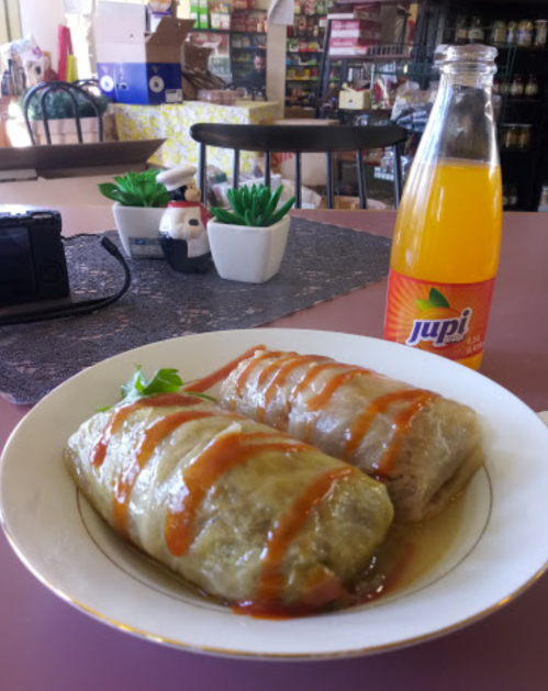 A plate of stuffed cabbage rolls drizzled with sauce, accompanied by a bottle of Jupi orange soda.