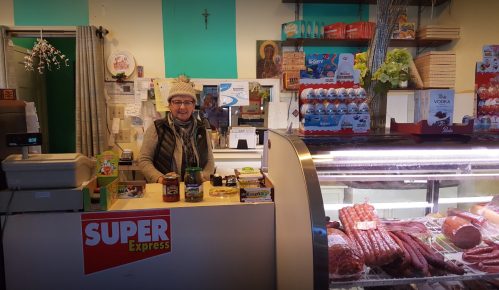 A smiling woman stands behind a counter in a small grocery store, surrounded by various food items and products.