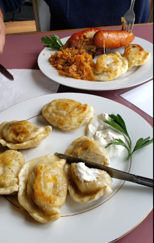 Two plates of food: one with pierogi and sour cream, the other with sausage and rice, garnished with parsley.