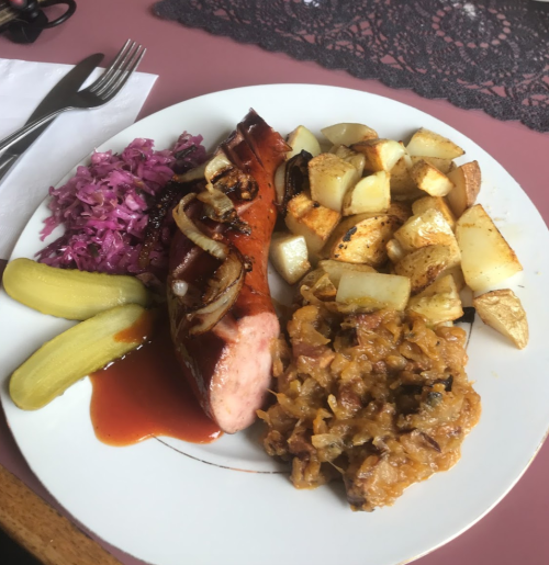 A plate with a grilled sausage topped with onions, roasted potatoes, sauerkraut, pickles, and coleslaw.