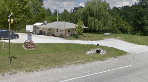 A small roadside building with a gravel driveway, surrounded by greenery and a picnic area.