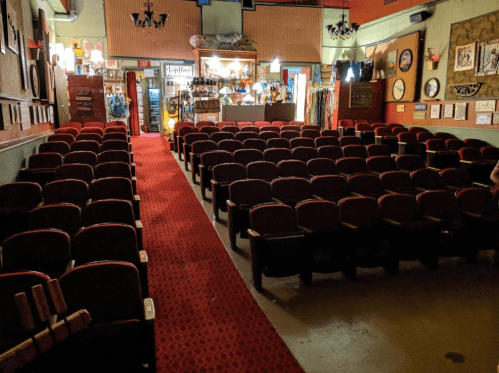 Interior of a vintage theater with red carpet and rows of red seats, leading to a brightly lit concession area.