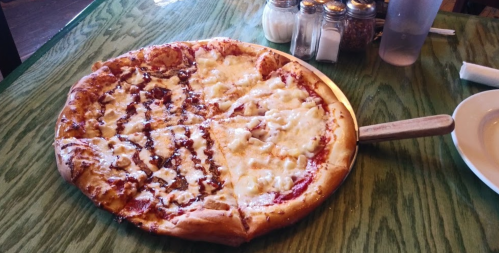 A large pizza with cheese and barbecue sauce, served on a wooden table with condiments in the background.