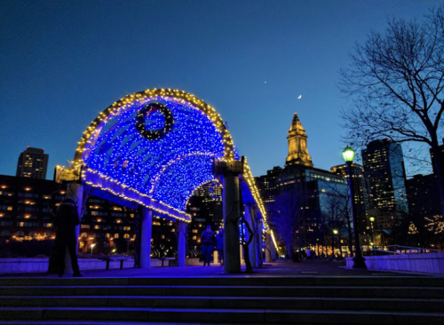 A lit archway with blue lights at dusk, framed by city buildings and a crescent moon in the sky.