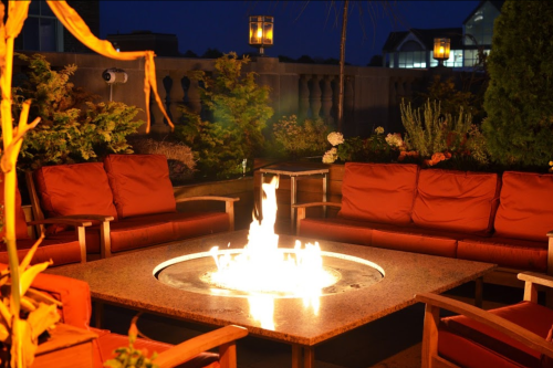 Cozy outdoor seating area with orange cushions around a circular fire pit, illuminated at night.