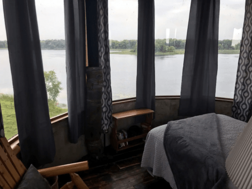 Cozy interior with a bed and wooden chair, featuring large windows overlooking a calm river and green landscape.