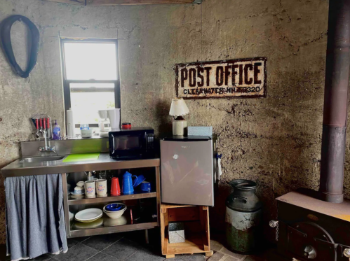 A rustic kitchen with a metal shelf, microwave, fridge, and a vintage "Post Office" sign on the wall.