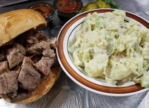 A sandwich filled with grilled meat next to a bowl of creamy potato salad, accompanied by small containers of sauce.