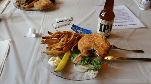 A plate with a sesame seed burger, fries, salad, a pickle, and a bottle of IPA beer on a table.