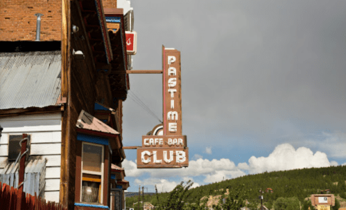 Sign for "Pastime Cafe-Bar Club" hanging from a building, with a cloudy sky and green hills in the background.