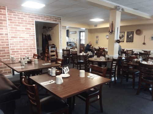 A cozy restaurant interior with wooden tables, chairs, and a brick wall, featuring a few diners and table settings.