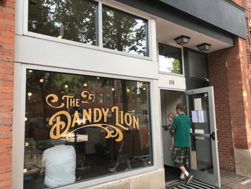 A storefront with the sign "The Dandy Lion," featuring large windows and a person entering with a child.