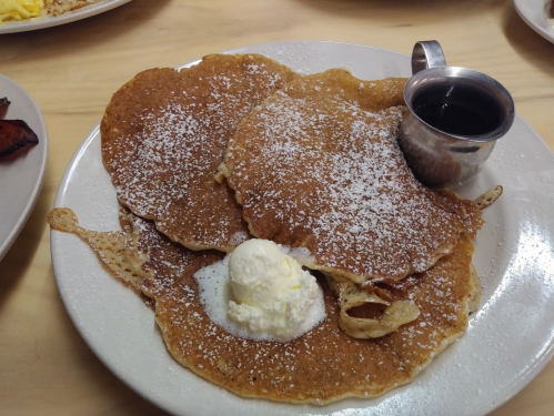 A plate of three large pancakes dusted with powdered sugar, topped with a scoop of butter and a small syrup cup.