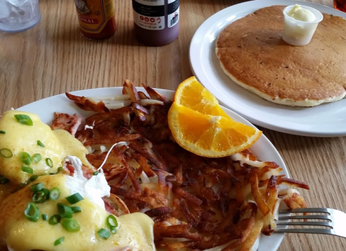 A plate of eggs Benedict with hash browns and orange slices, alongside a pancake with butter on a wooden table.